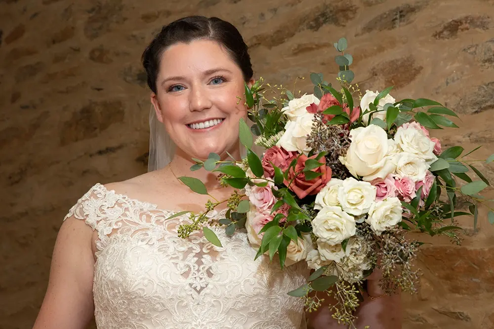 Smiling bride in white lace dress holds large floral bouquet.