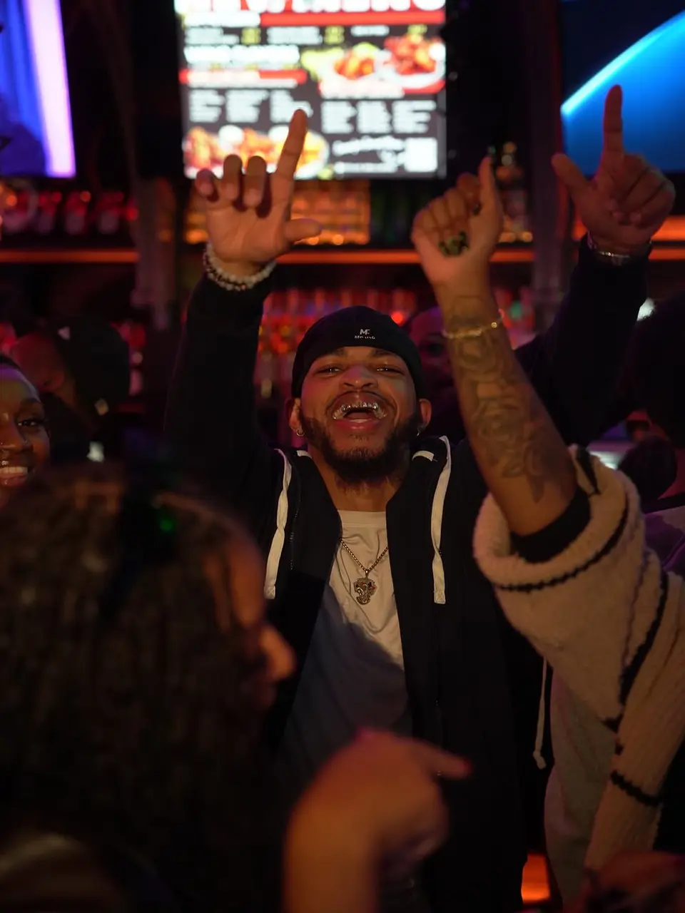 Man with arms raised, smiling, next to friends in a bar setting