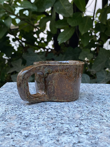 Side view of a small, handmade ceramic cup with a thick handle, placed on a granite surface against leafy greenery.
