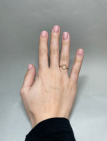 Hand with pink nails wearing a minimal brass ring with a central circle on the ring finger, against a gray background.