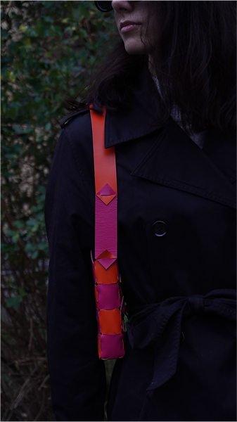Woman in black coat with a shoulder strap featuring pink and orange geometric pattern, wearing sunglasses outdoors.