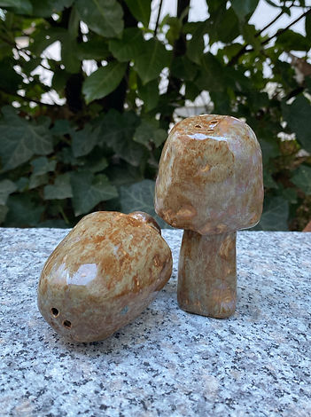 Two ceramic mushroom sculptures with earthy tones sit on a stone surface, surrounded by green ivy leaves.