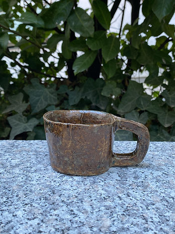A small, rustic ceramic cup with a large handle sits on a granite surface, backed by dense green foliage.