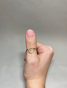 Thumb wearing a brass ring with a loop and wave design, hand held upright against a light gray background.