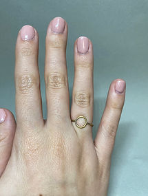 Close-up of hand with pink nails wearing a brass ring with a central circle on the ring finger, shown against a gray backgrou