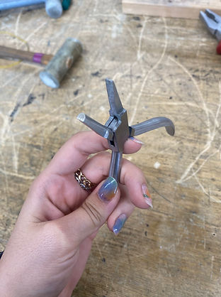 Hand with colorful nails holding a multi-functional jewelry tool above a wooden workbench.