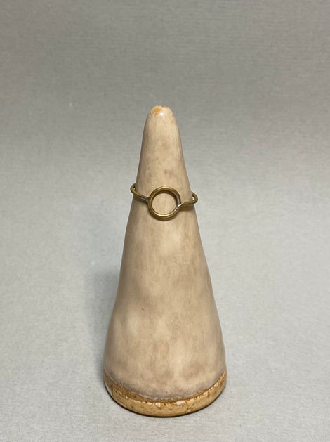 Brass ring with a central circular element displayed on a ceramic cone stand against a soft gray background.