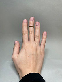 Hand with pink nails wearing a thin brass ring on the middle finger, shown against a gray background.