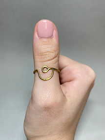 Close-up of thumb wearing a brass ring with a loop and curved wave design, shown against a neutral gray background.