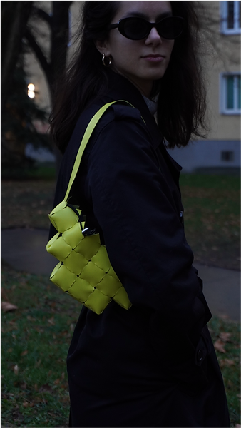 Woman in black coat and sunglasses carries a small bright yellow woven bag over her shoulder in an outdoor setting.