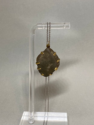 Pendant necklace with an irregular dark stone held by brass prongs, hanging on a silver chain and clear stand.