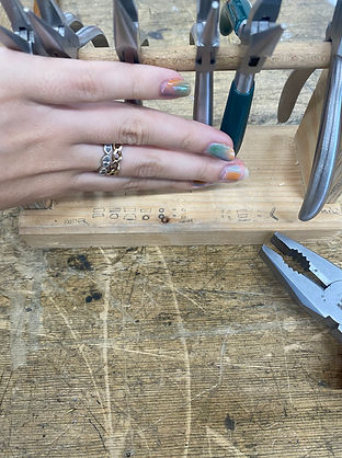 Hand with colorful nails and stacked rings resting near jewelry pliers on a wooden workbench.