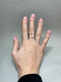 Hand with pink nails wearing an open brass ring with curled ends on the middle finger, shown against a gray background.
