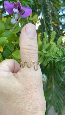 Hand with brass wave-shaped ring on thumb, held up near a purple flower and green leaves in natural light.