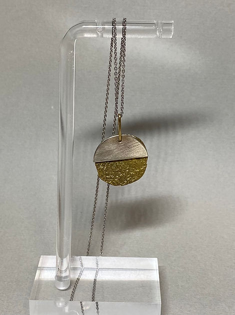 Pendant necklace with a circular charm, half brushed silver and half textured gold, displayed on a clear stand.