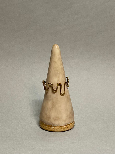 Brass wave-pattern ring displayed on a ceramic cone stand against a neutral gray background.