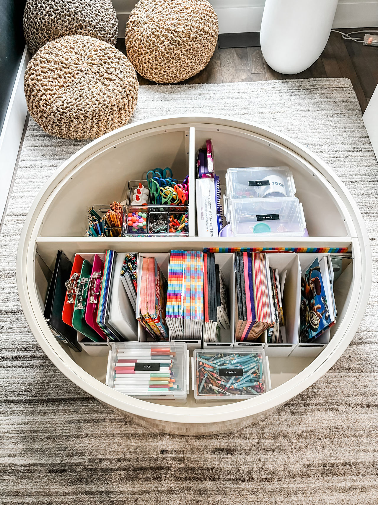 Round table with organized crafts: scissors, markers, and notebooks in compartments. Beige poufs on a rug background.
