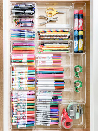 Organized drawer with colorful markers, pencils, scissors, glue, and tape in clear compartments. Neat and colorful arrangement.
