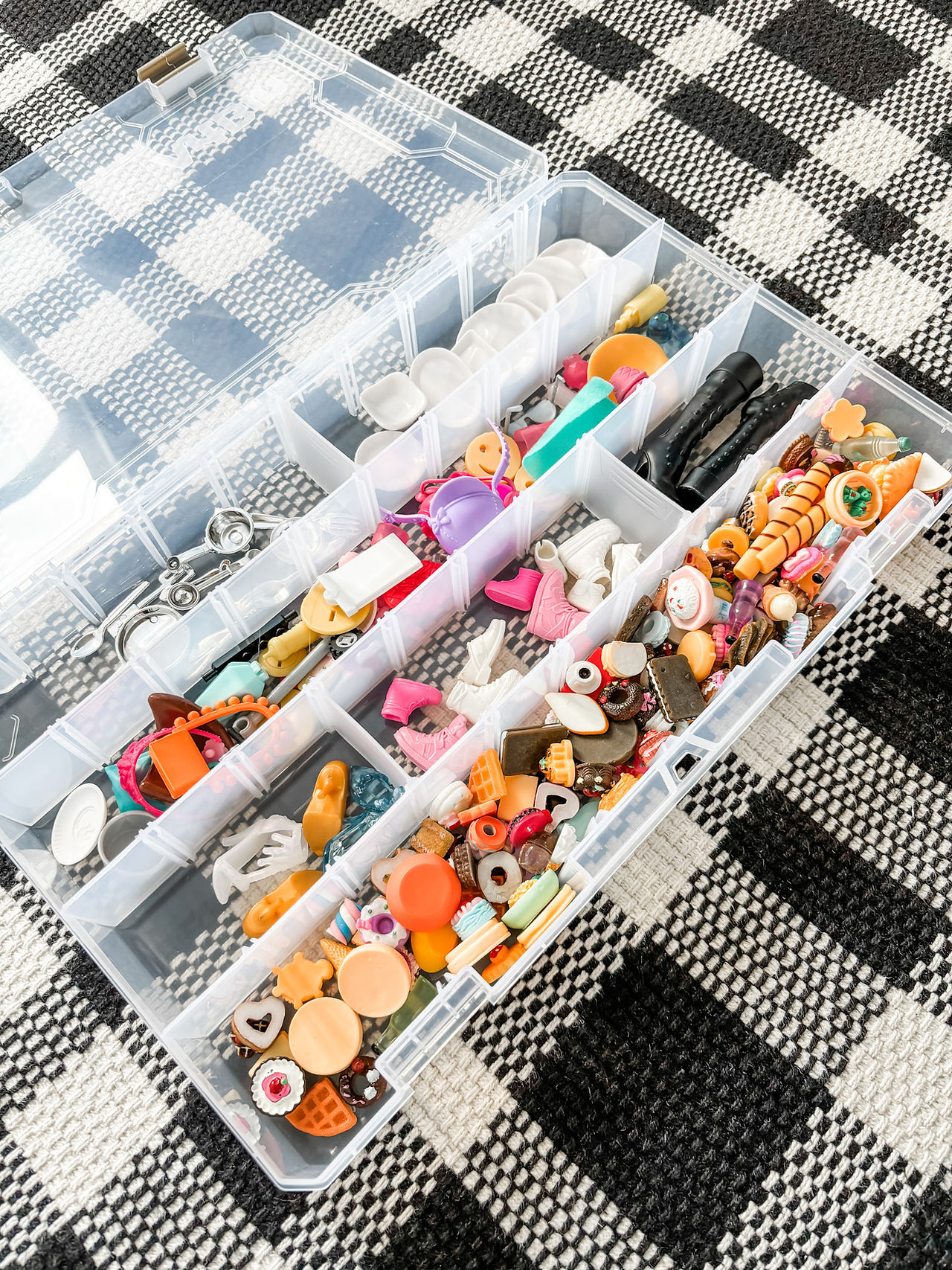 Clear organizer box with various colorful small trinkets on a black-and-white checkered surface. Items include charms, toys, and buttons.