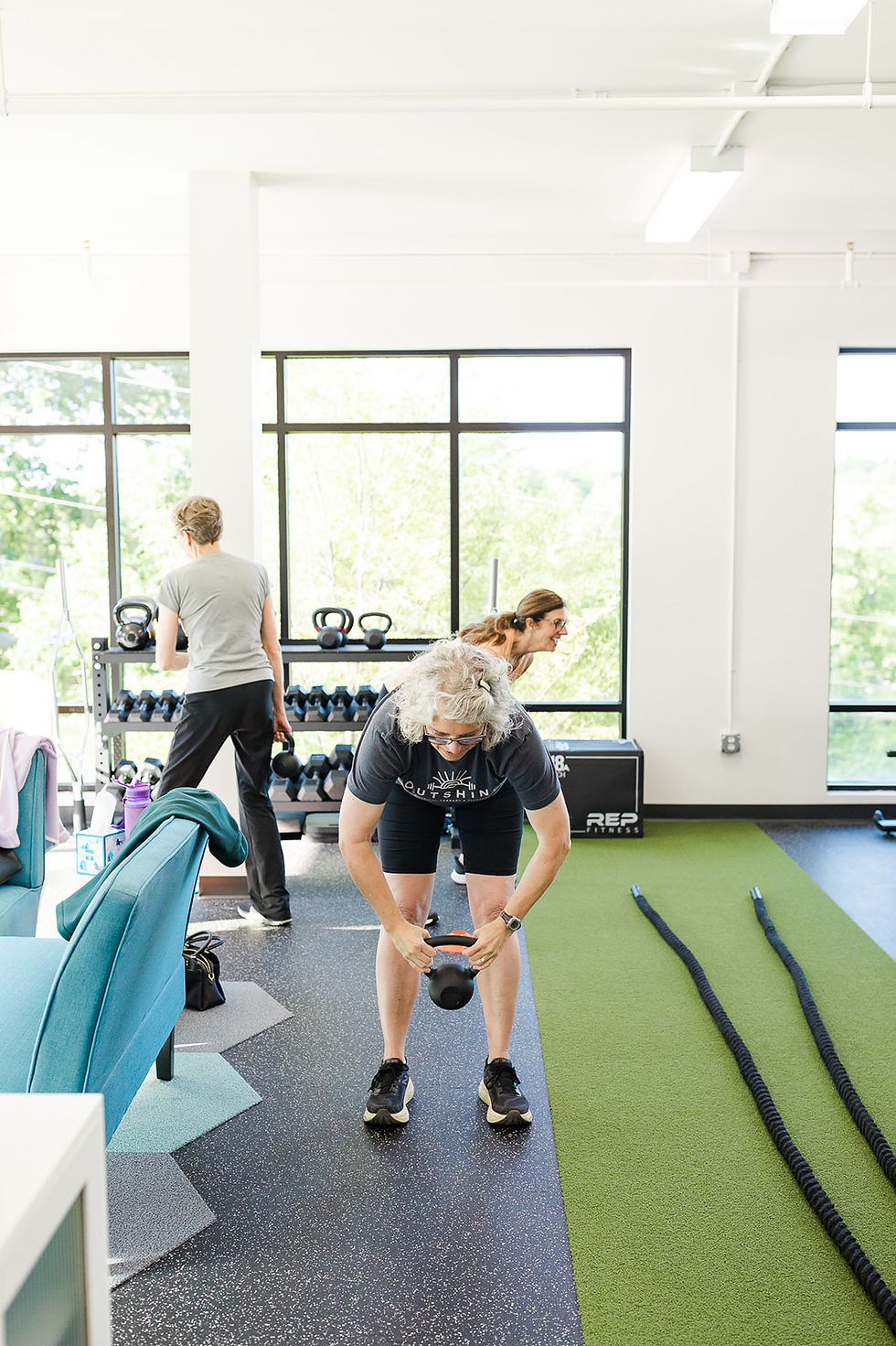 Three people exercising in a bright gym; one lifts a kettlebell, others near dumbbell rack. Green turf and exercise ropes visible.