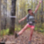 A hiker poses with her leg and arms out, smiling on a trail covered in fallen leaves. A sign nailed to a tree behind her marks the terminus of the trail