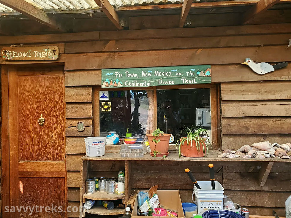 The wooden front door and side of a house with various plants and gardening tools on a table and bench in front of a window with a green sign that reads "Pie Town, New Mexico on the Continental Divide Trail"