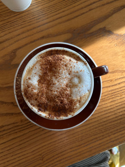 An top down photo looks down on a coffe cup sitting on a small plate. The plate is a dark purple color. The handle of the cup is the same color as the plate. The coffee inside the cup is topped with foam and a sprinkle of powder such as cinnamon. The cup and plate sit atop a wood table.