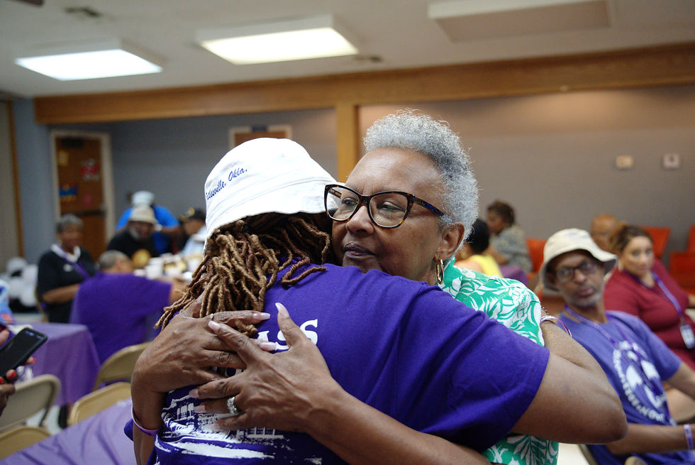 Two women share a warm hug during an indoor gathering, one wearing a purple Douglassaires T-shirt and white hat, the other wearing glasses and a green patterned blouse. People are seated at tables in the background.