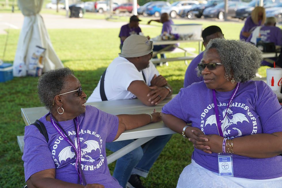 Two women wearing purple Douglassaires reunion T-shirts sit at a picnic table outdoors, talking and smiling. Other attendees in similar shirts are visible in the background under a large event tent.