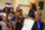 A smiling young woman with long braids holds a large scholarship check while standing beside another woman in a floral dress. Several people stand in the background, including a man in a suit and tie. Purple and white balloon decorations are visible, with text overlay reading 'Carrying forward a legacy built on resilience, excellence, and pride.'