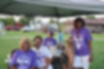 Five smiling adults gathered under a canopy at the 2023 Douglassaires reunion, with three wearing purple reunion T-shirts and one wearing a white reunion T-shirt. They are seated at a picnic table with drinks, while a grassy park and vehicles are visible in the background.