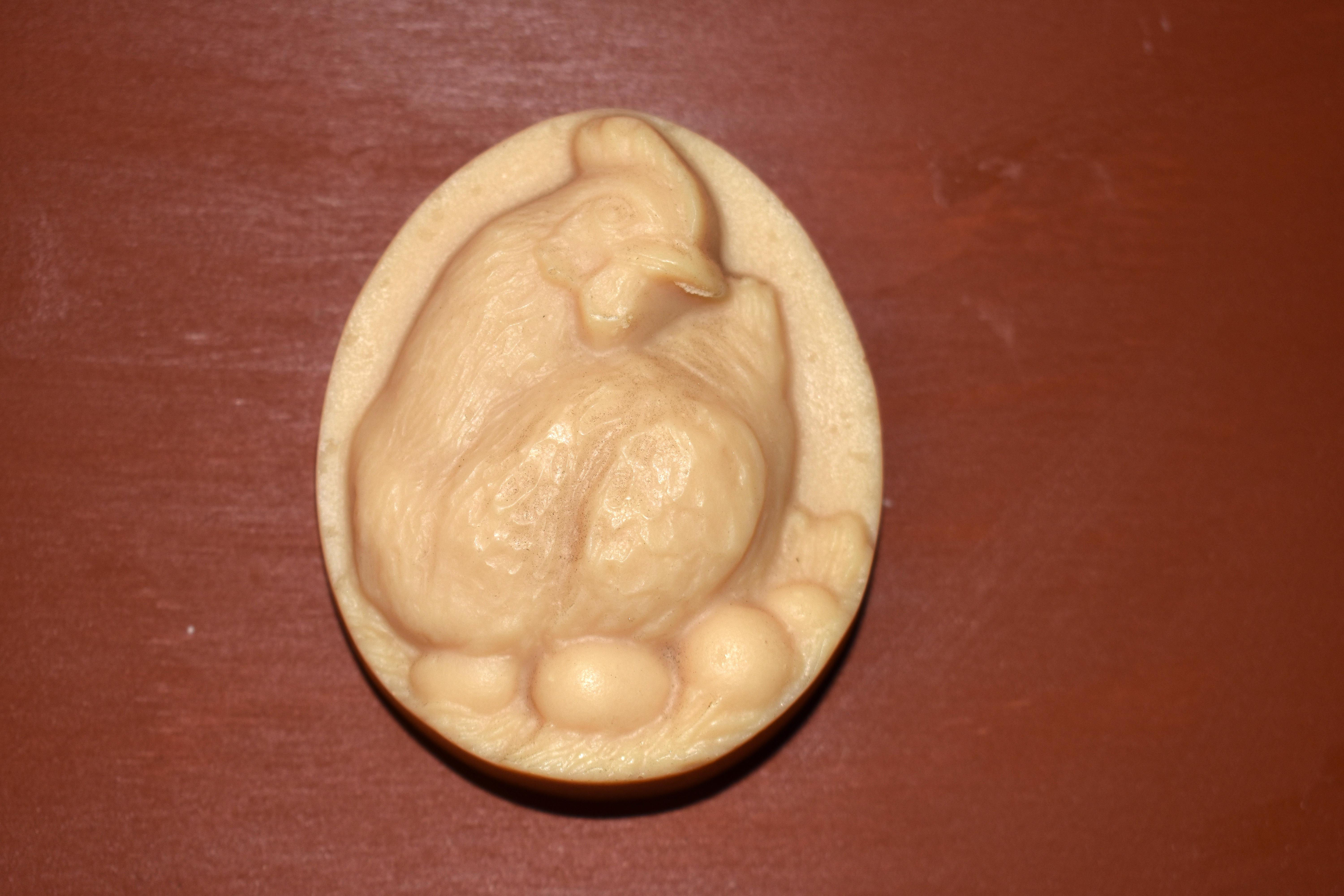 An egg shaped soap, about the size of an adult palm, with a raised image of a hen resting on five eggs in a nest.
