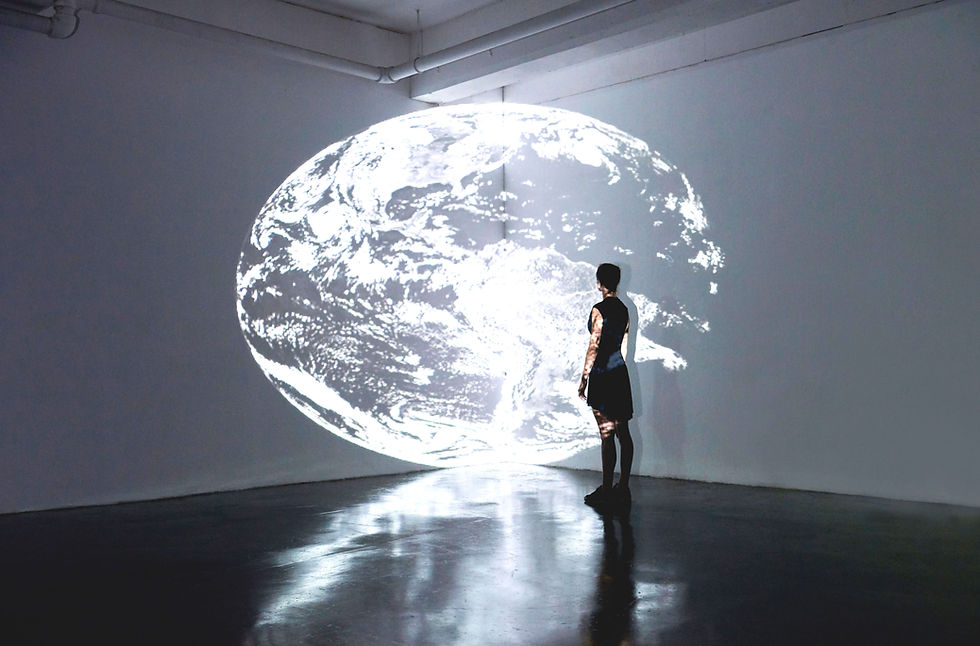 A person in a dark room gazes at a large projected image of Earth on a wall. The scene is minimalistic, with a reflective floor.