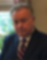 Stephen Akin in a suit with a striped tie stands indoors, looking at the camera. Background shows a window with greenery outside and curtains.