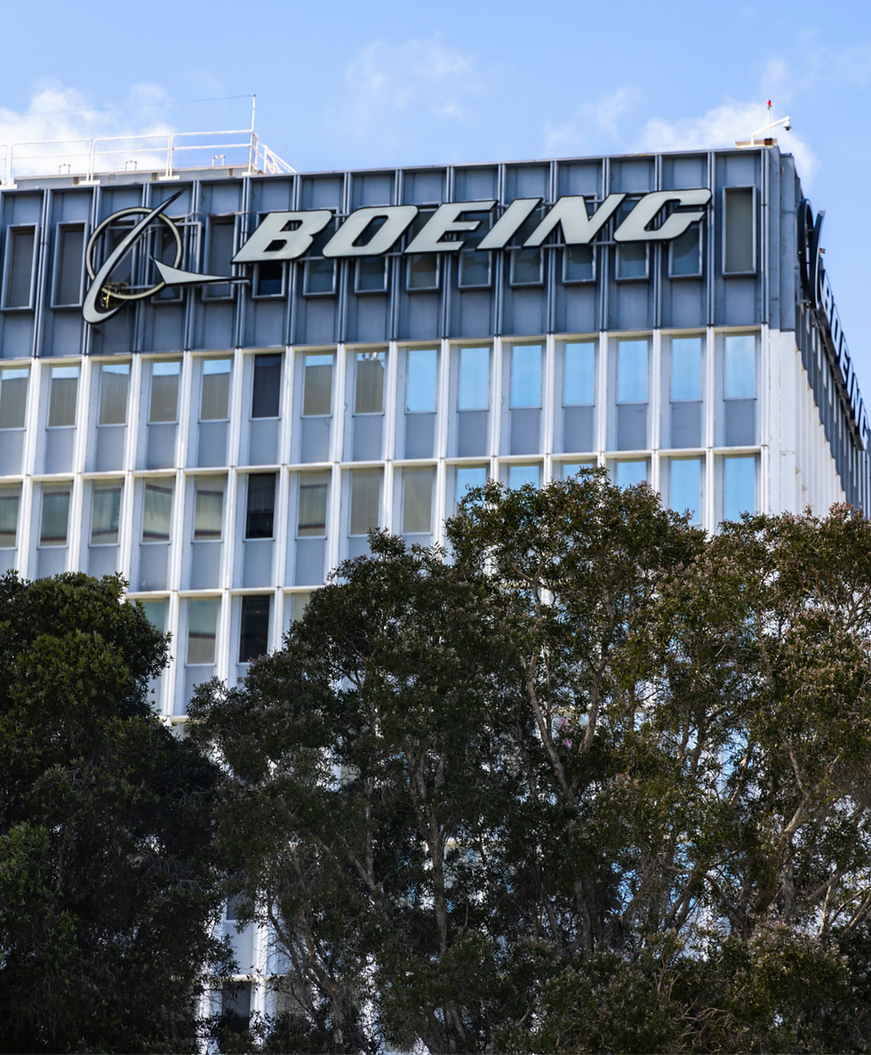 Office building facade with "Boeing" logo at top. Large windows and trees in foreground, set against a partly cloudy sky.