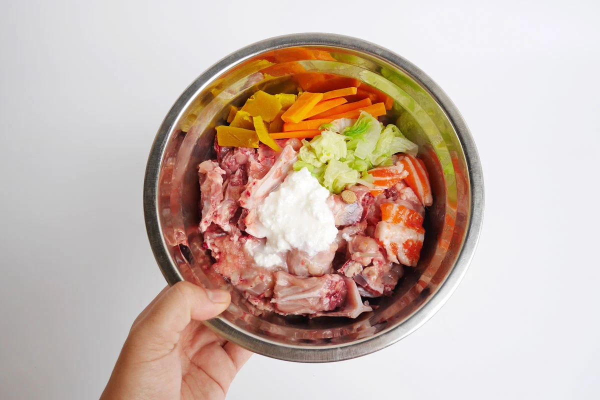 Pet food bowl with a variety of fresh vegetables, raw meat, and yogurt