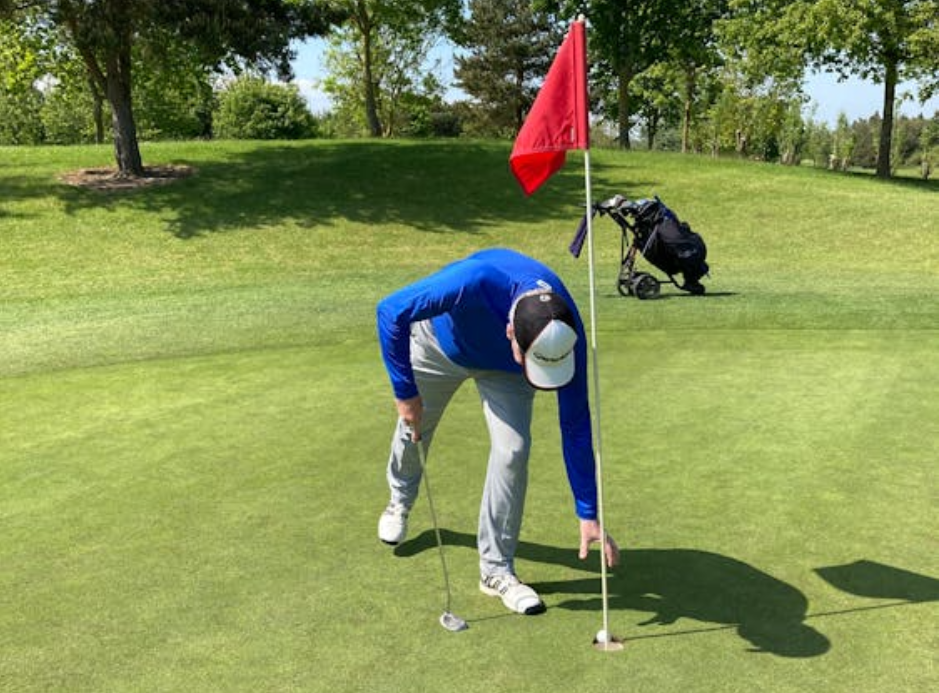 Golfer in a blue long-sleeve shirt and gray pants bending down to retrieve a ball from the hole on a sunny green, with a red flagstick and golf bag visible in the background.