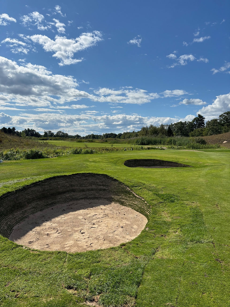 Sunny golf course with a deep sand bunker in the foreground and another bunker further back, surrounded by well-kept green grass, trees, and open blue sky with scattered clouds.