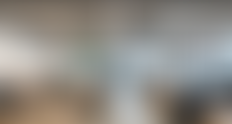 Modern open-plan office with natural light, minimalist wooden desks, exposed concrete ceiling, and hanging plants. Employees work at computers in a collaborative, eco-friendly workspace with industrial lighting.