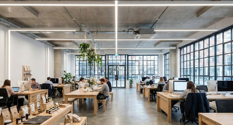 Modern open-plan office with natural light, minimalist wooden desks, exposed concrete ceiling, and hanging plants. Employees work at computers in a collaborative, eco-friendly workspace with industrial lighting.