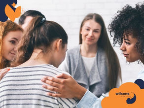 A group of women in a supportive circle, one comforting another with a hand on her shoulder. They look empathetic. Wilsons Health logo present.