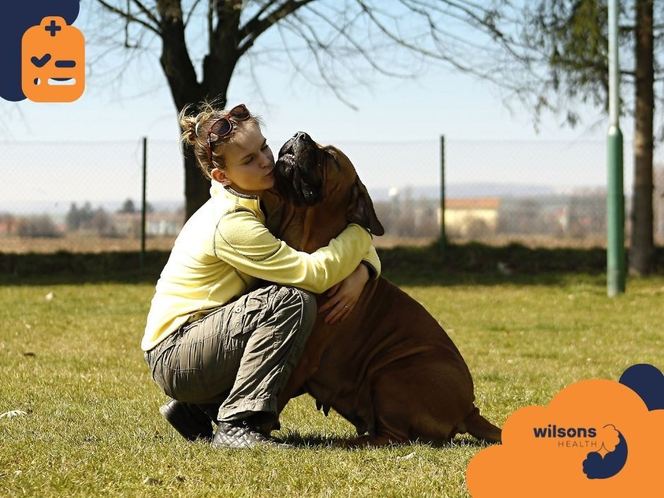 Person hugging a large brown dog on grass, showing affection. Background has trees and a fence. Logo text: "wilsons health."