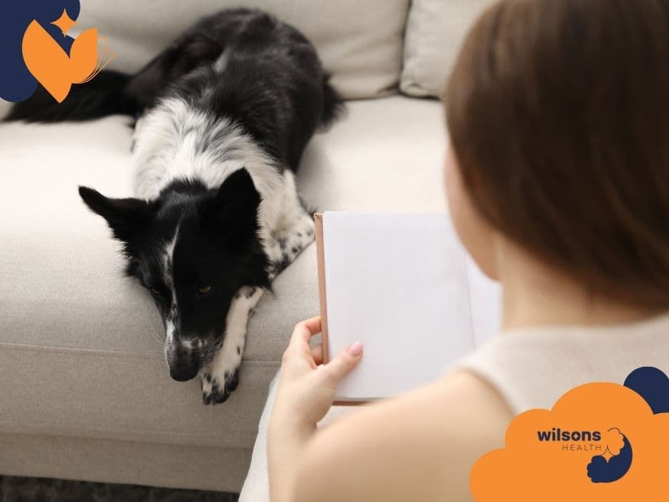 Person reading a book on a beige couch with a black and white dog resting nearby. "Wilsons Health" logo in the corner. Calm atmosphere.