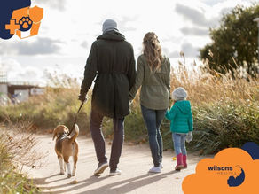 Family walking a beagle on a path through tall grass under a cloudy sky. They're dressed warmly. "Wilsons Health" logo visible.