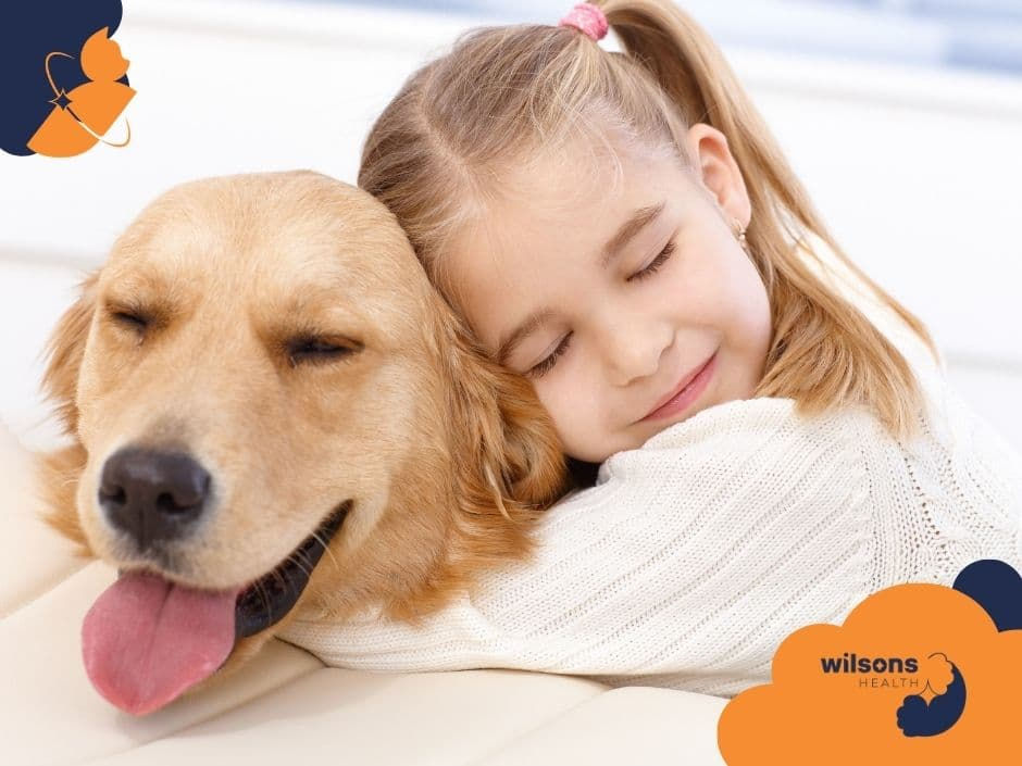 Girl hugging a golden retriever on a white sofa, both with eyes closed. The scene conveys warmth and comfort. Wilsons Health logo visible.