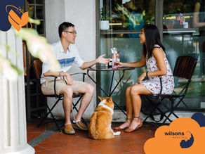 A couple sits at an outdoor café table, chatting. A brown and white dog sits nearby. Visible logo: "Wilson's Health." Warm, casual setting.