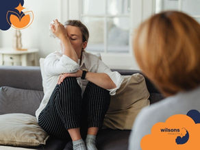 A person sits on a sofa, wiping tears, looking distressed. Another person is blurred in the foreground. Text reads Wilsons Health.