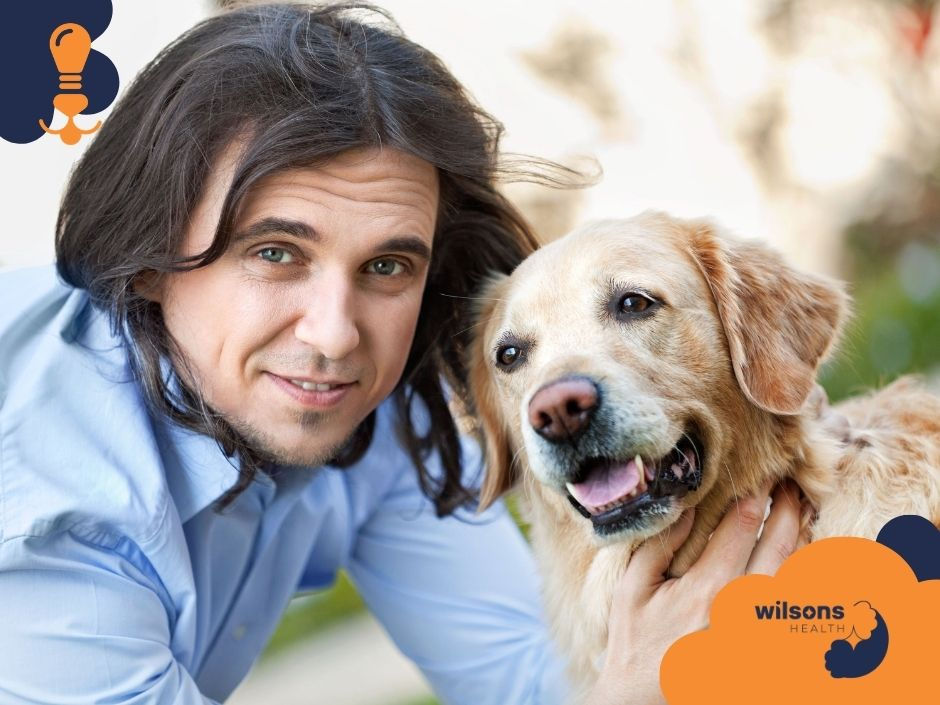 Man in a blue shirt smiles, hugging a golden retriever. The logo "wilsons HEALTH" is in the corner. Outdoor setting, bright and cheerful mood.