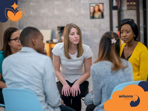 A diverse group of five people sits in a circle, engaged in a discussion. The background shows bookshelves and a gray wall. Text: wilsons health.