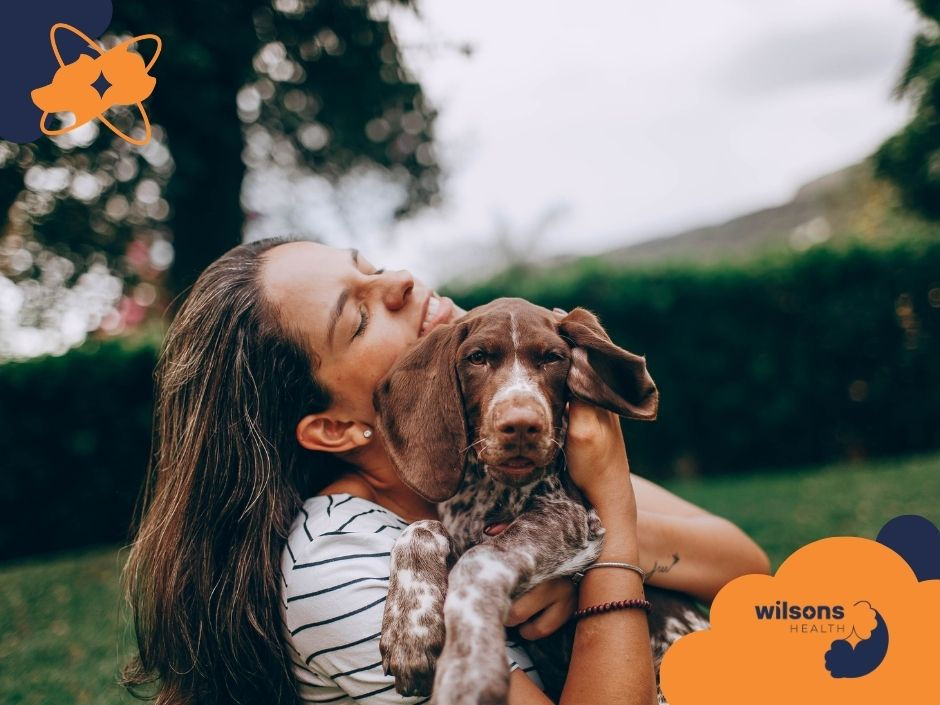 Woman smiling, cuddling a brown puppy outdoors. Greenery in the background. Logo with "wilsons HEALTH" is visible. Warm, joyful mood.